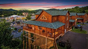 A wooden cabin with a balcony, surrounded by a parking area and a view of a town and mountains.