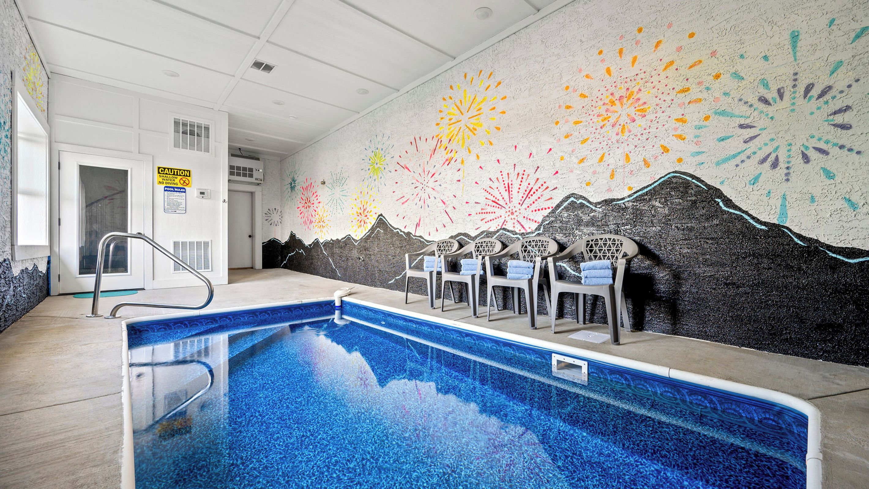 An indoor swimming pool with a blue tiled floor, a metal pool ladder, and a wall mural with mountain and fireworks designs.