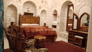Una habitación con una cama grande, cabecera de madera y un espejo. Hay dos mesitas de noche con lámparas y un aparador con espejo. La habitación tiene arcos de piedra en las paredes.