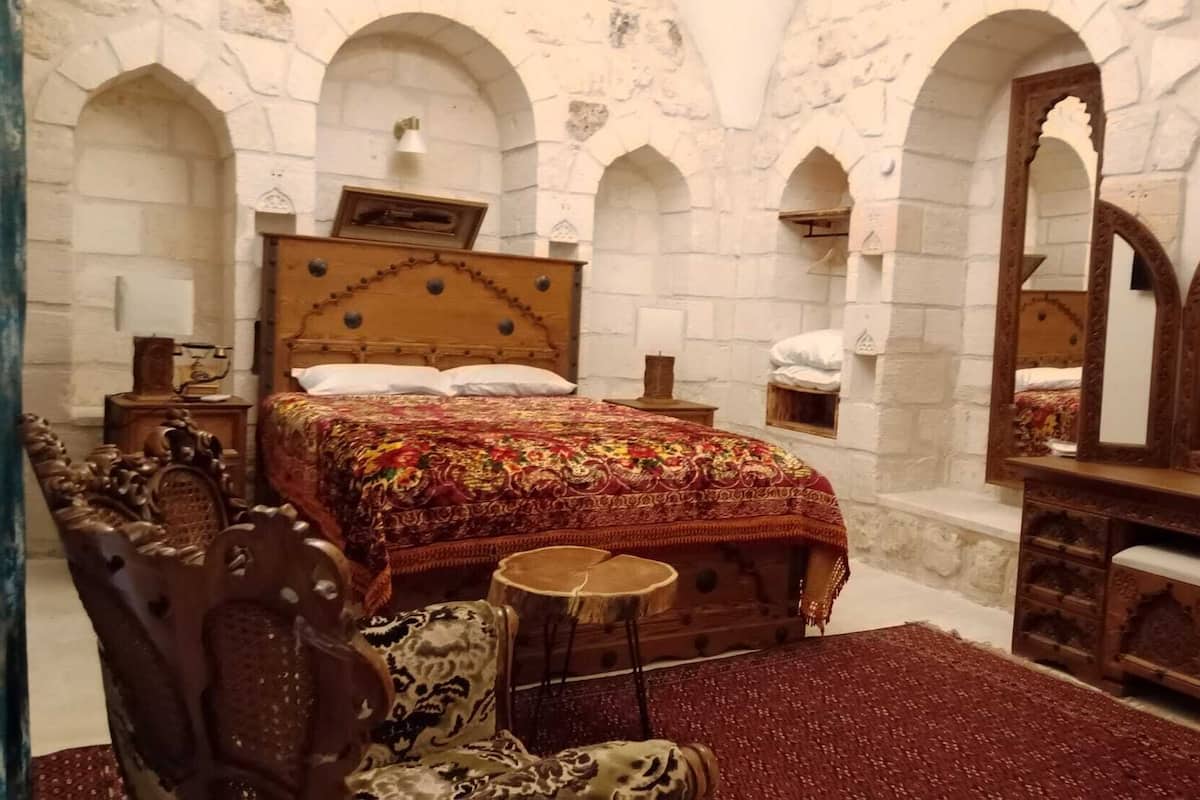 Ein Zimmer mit einem großen Bett, einem Holzkopfende und einem Spiegel. Zwei Nachttische mit Lampen sowie eine Kommode mit Spiegel gehören zur Einrichtung. Die Wände sind mit Steinbögen versehen.