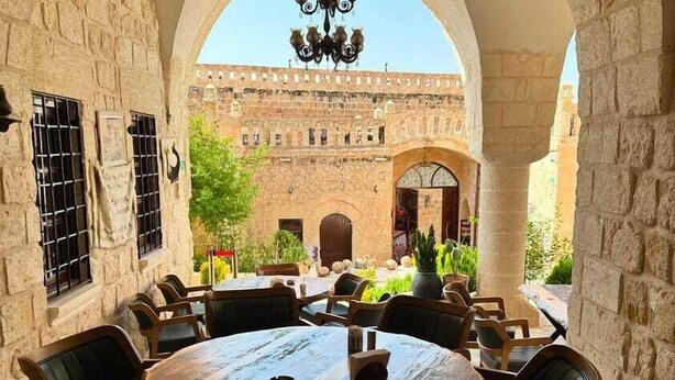 Un área de comedor al aire libre con una mesa y sillas de madera redondas, paredes de piedra y vistas a un edificio histórico al fondo.