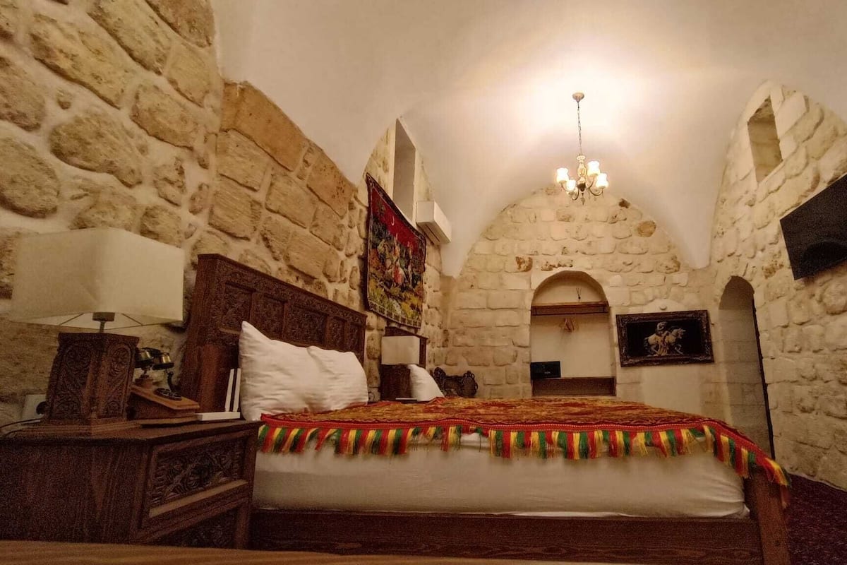 Ein Schlafzimmer mit Steinwänden, einem hölzernen Bett, einer Chandelierleuchtre und einer kleinen, gewölbten Tür.