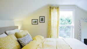 Een slaapkamer met een bed, kussens, een raam met gordijnen en ingelijste foto’s aan de muur.
