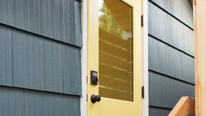 Een houten trap die leidt naar een gele deur met een ruitje, voor een grijze buitenmuur.