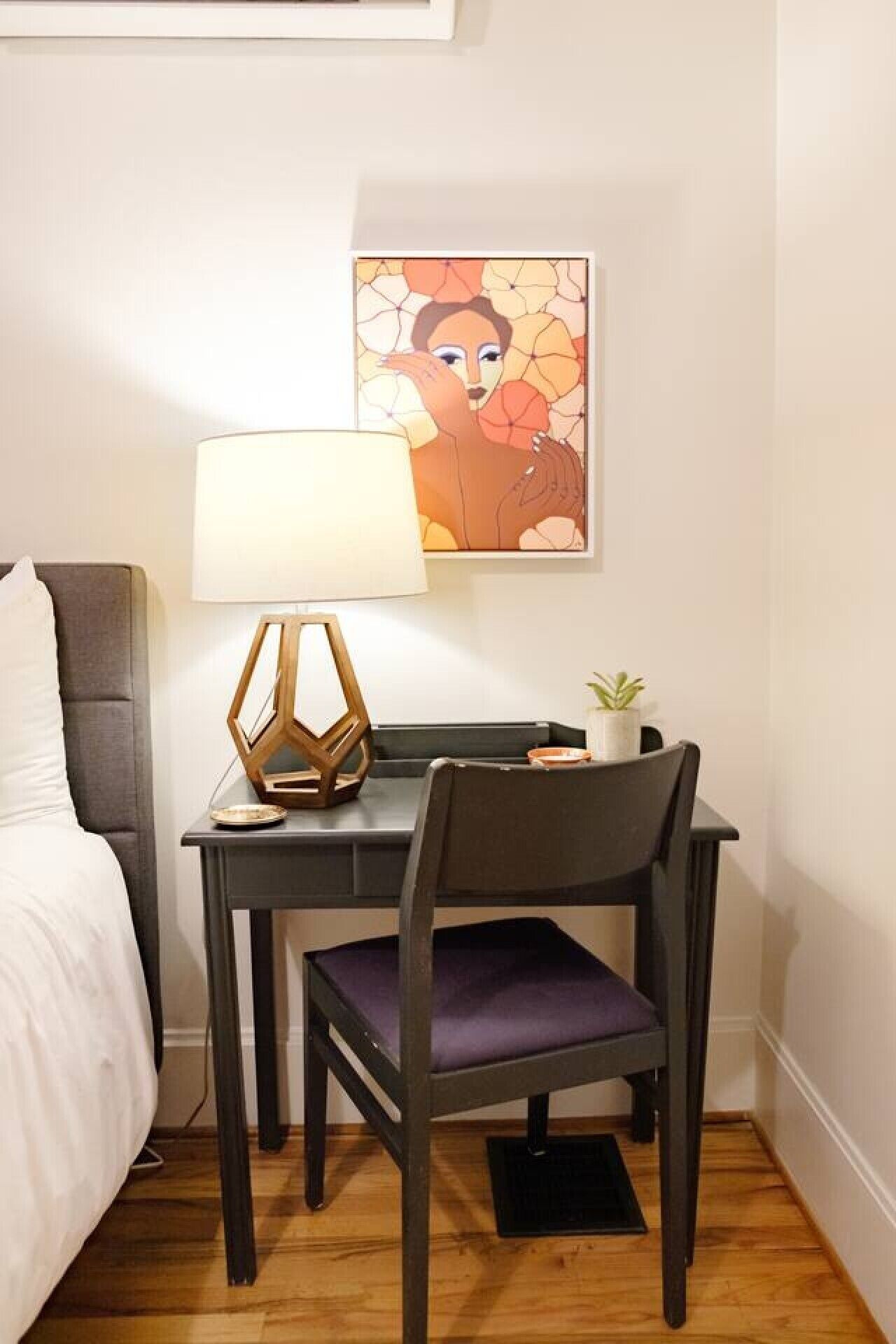 A small, neatly arranged hotel room with a bed, a desk with a lamp, a chair, and a wall-mounted painting.