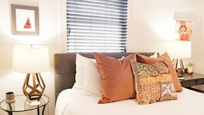 A hotel room with a bed, two bedside tables with lamps, a window with blinds, and a framed picture on the wall.