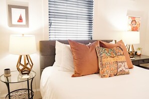 A hotel room with a bed, two bedside tables with lamps, a window with blinds, and a framed picture on the wall.