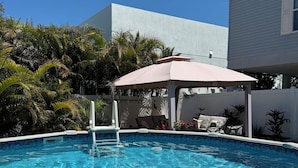 Una piscina con muelle blanco, zona de estar techada con cojines y cerca blanca.