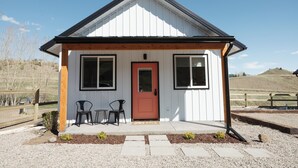 A small, single-story house with a red door, two windows on the front, and a porch with two chairs and a table. - NEW Farm Stay Cabin close to Livingston, Yellowstone, Bozeman, fishing, hiking (Livingston)