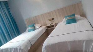 A hotel room with two single beds, each with a white bedspread and a teal pillow. There is a wooden headboard, a bedside table with a lamp, and a window with blue curtains. - Junting Hotel, Taohua Island (Zhoushan)