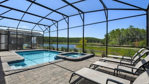 A covered outdoor swimming pool with lounge chairs and a hot tub, surrounded by a metal frame and glass walls, overlooking a lake and forest. - Lake View Cozy Home w/ Pool & Games Near Disney (Kissimmee)