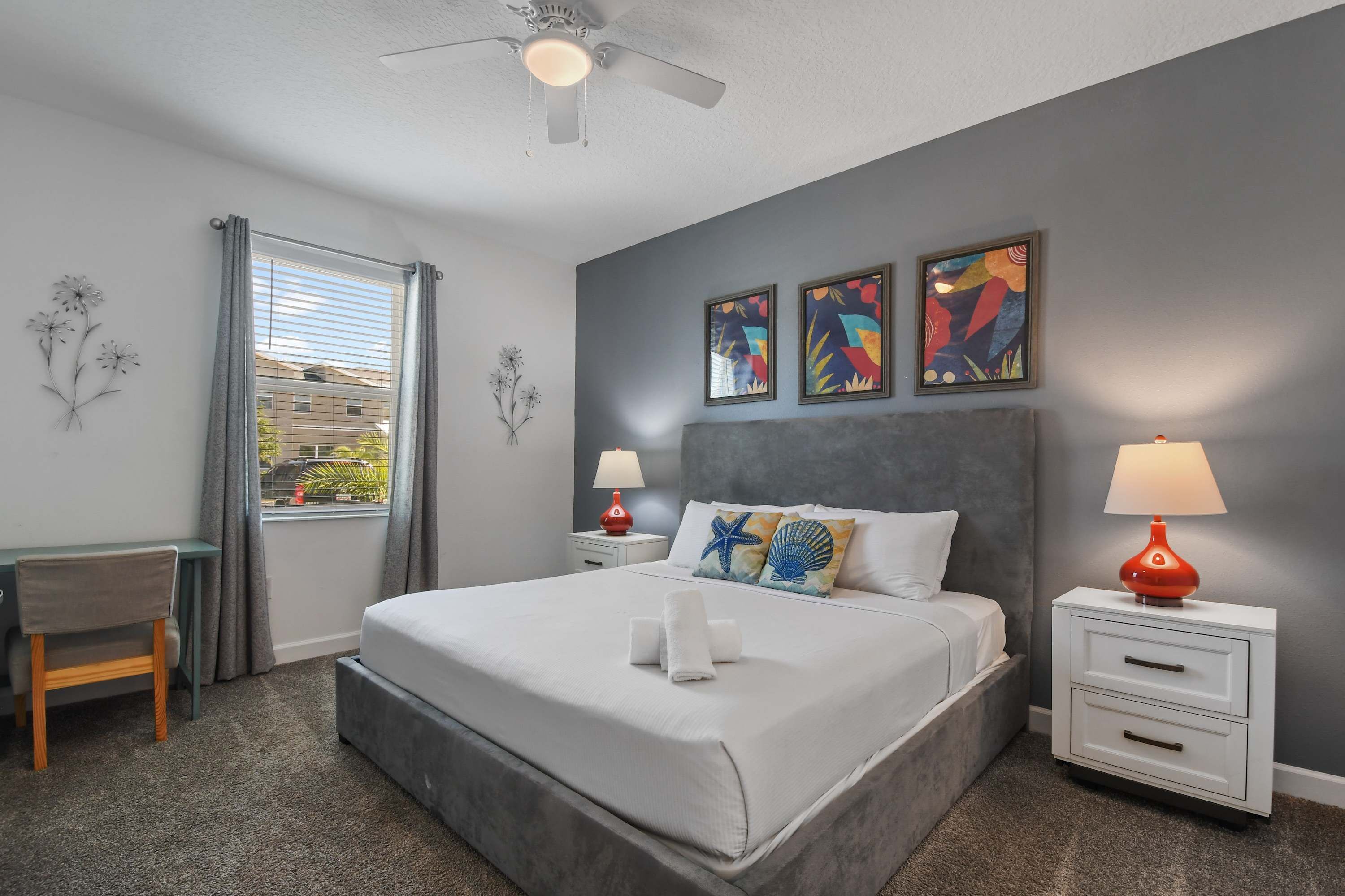 A bedroom with a bed, two bedside tables, a desk, a chair, a ceiling fan, and artwork on the wall.