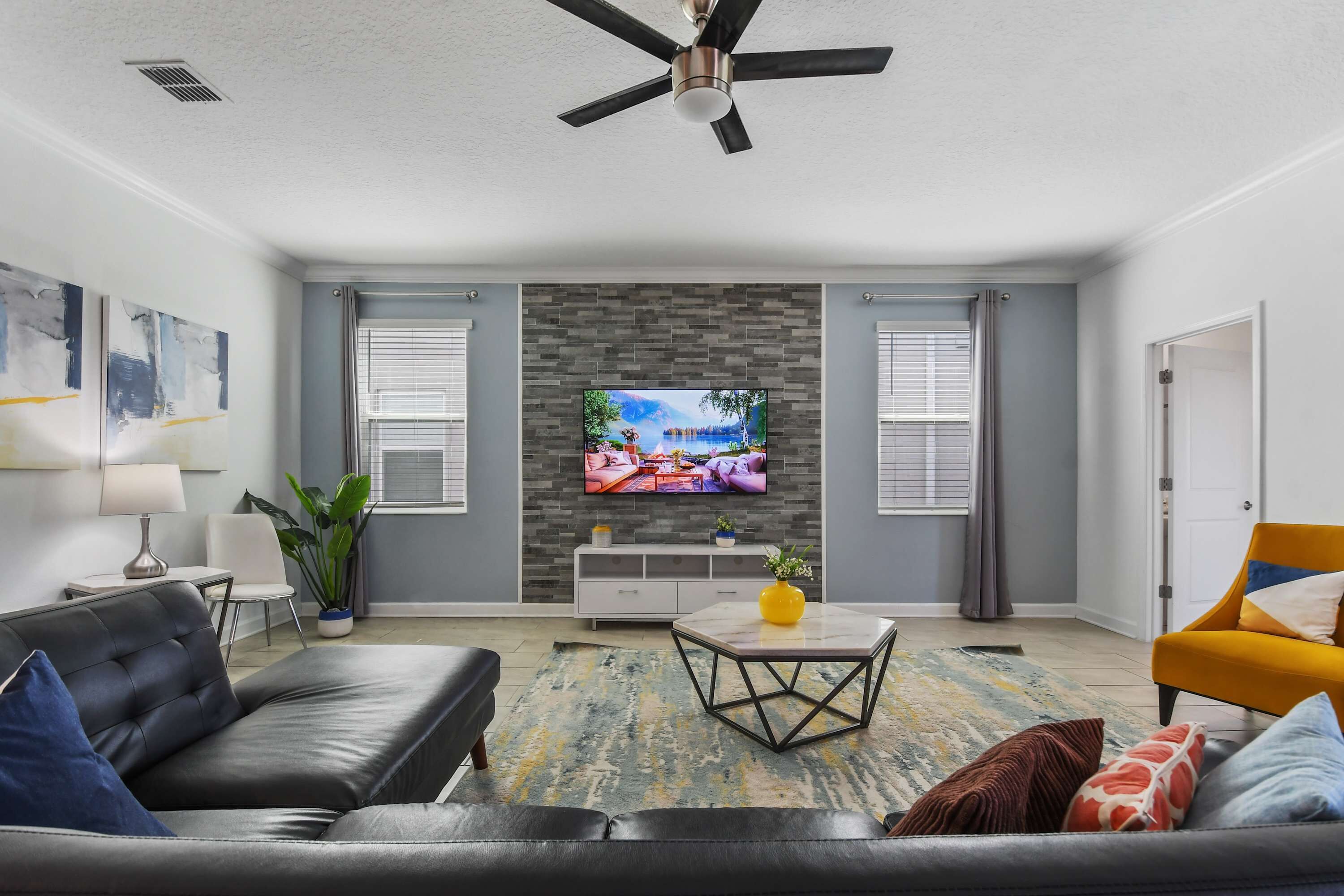 A modern living room with a grey sofa, a yellow armchair, a coffee table, and a flat-screen TV mounted on a brick wall.