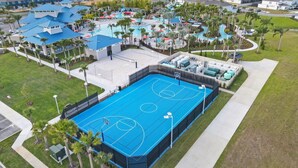 An aerial view of a resort with a large blue basketball court, multiple swimming pools, and palm trees.