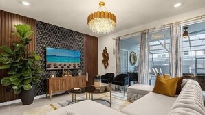 A modern living room with a large TV, comfortable seating, a chandelier, and a large window with curtains.