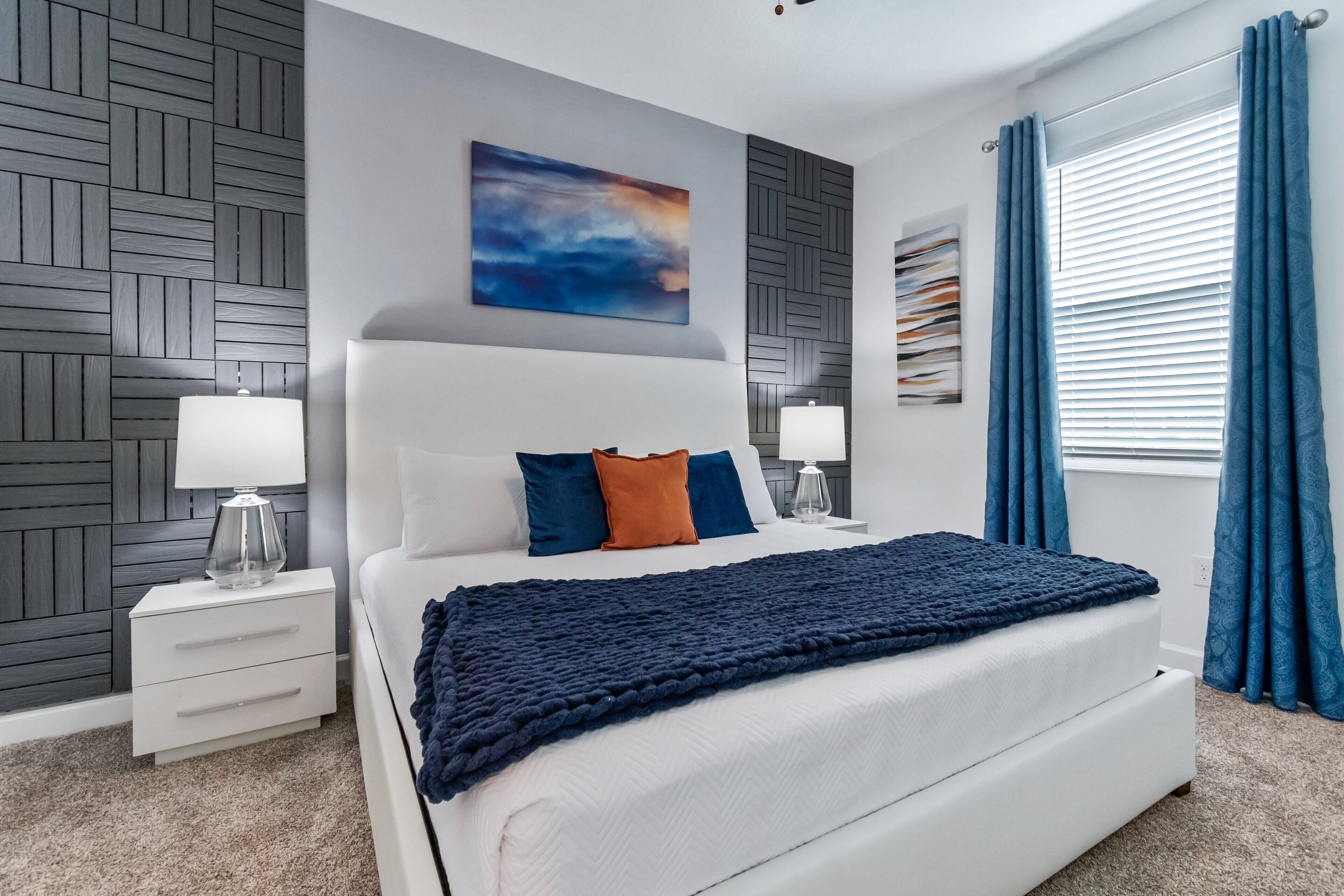 A modern bedroom with a large bed, two bedside tables with lamps, a wall-mounted artwork, and a window with curtains.