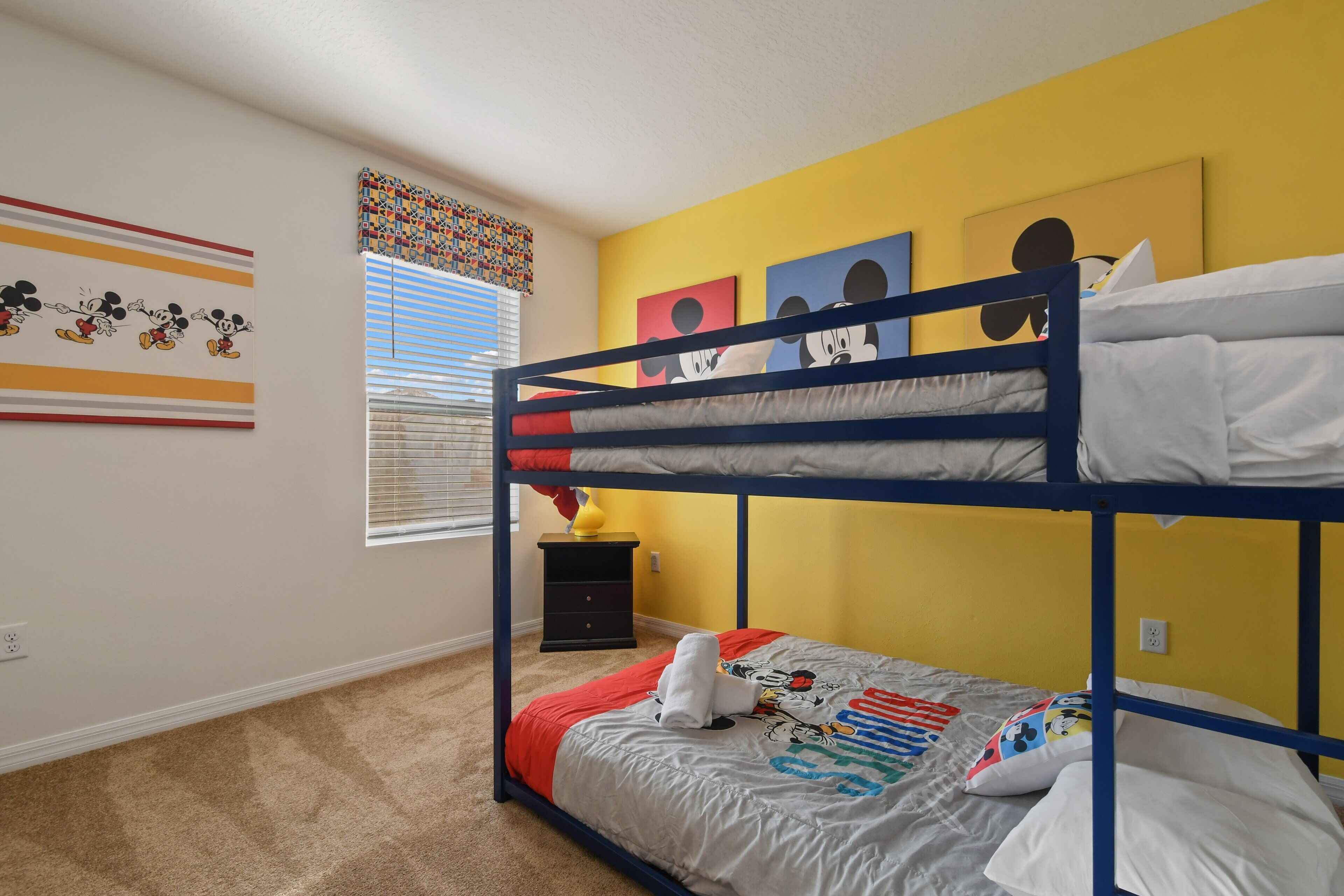 A bunk bed room with a yellow wall, a window with blinds, and Mickey Mouse themed decorations.