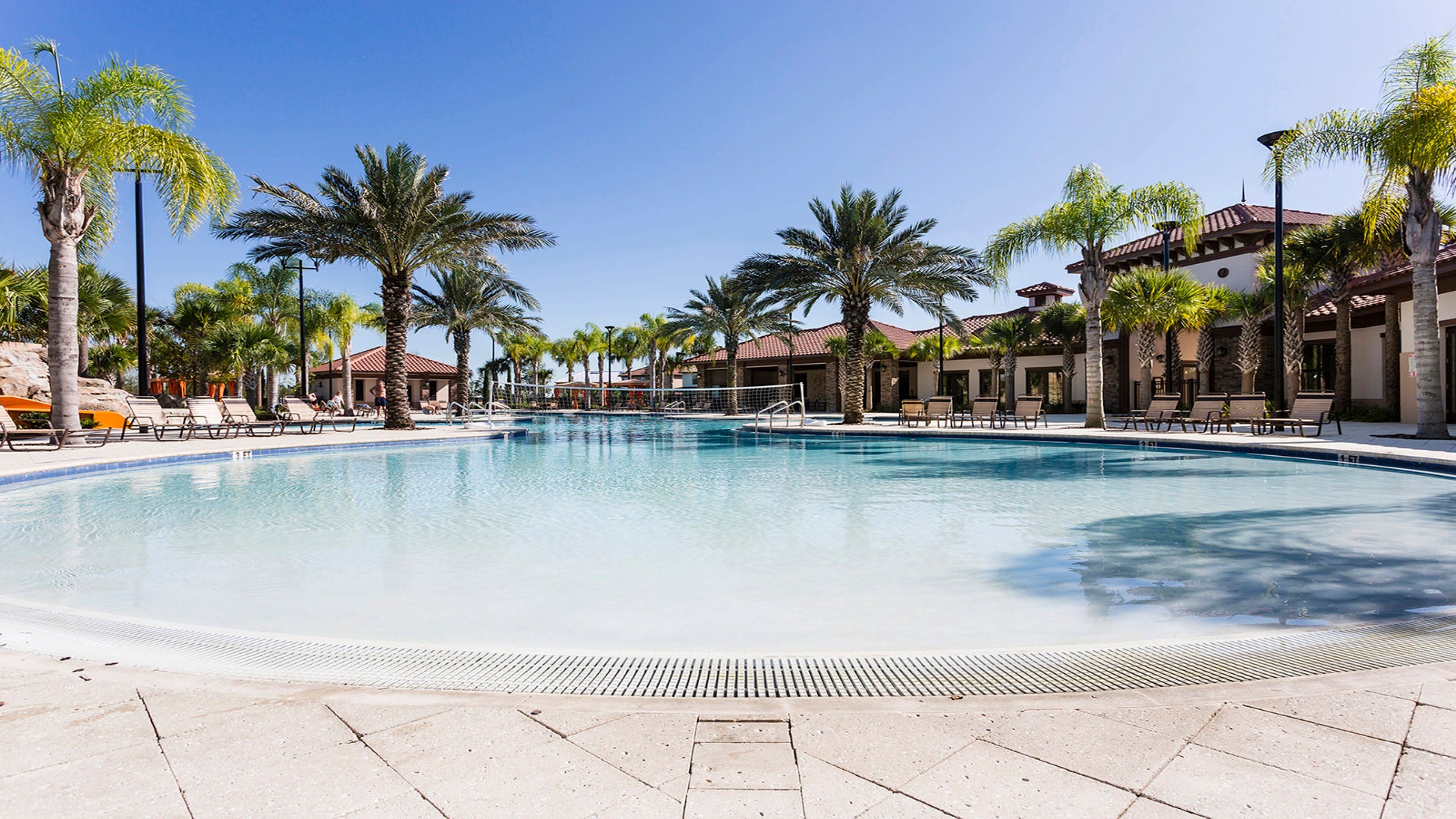 A large outdoor swimming pool surrounded by palm trees and lounge chairs, with a clear blue sky above.