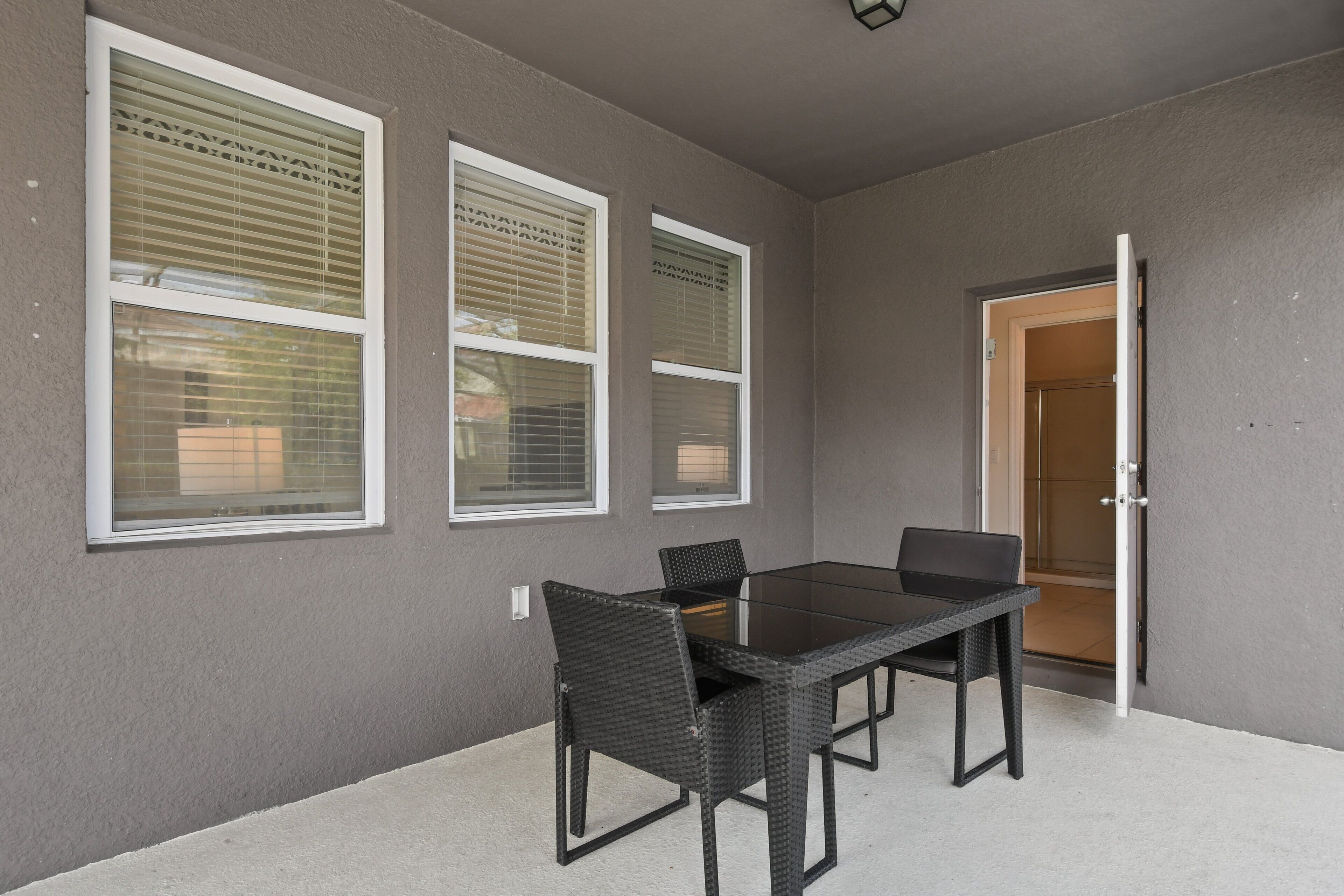 Une terrasse extérieure avec une table et des chaises, trois fenêtres équipées de stores et une porte donnant sur un espace abrité.