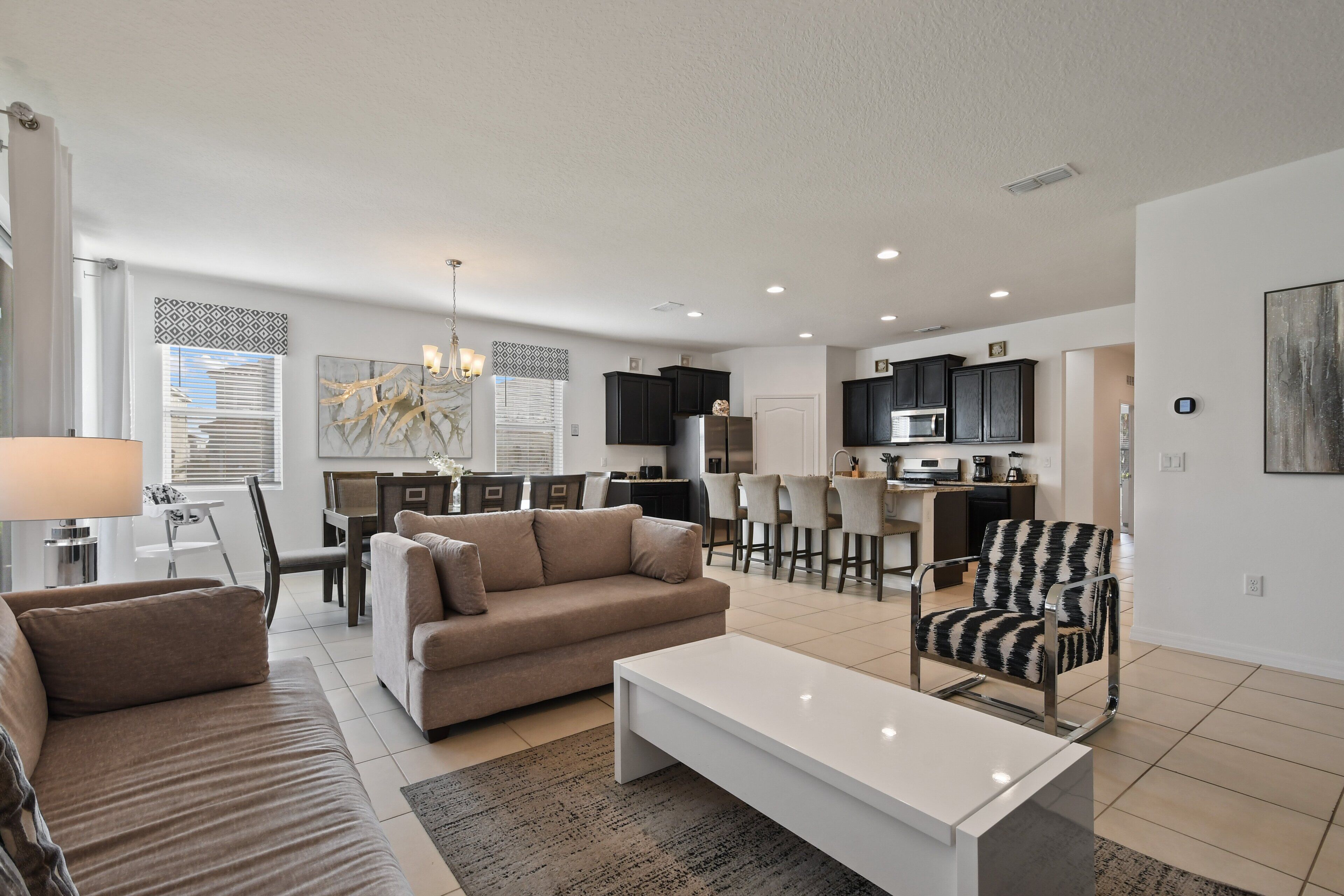 A modern living room with a sofa, coffee table, and patterned armchair. A dining area with a table and chairs is visible in the background.