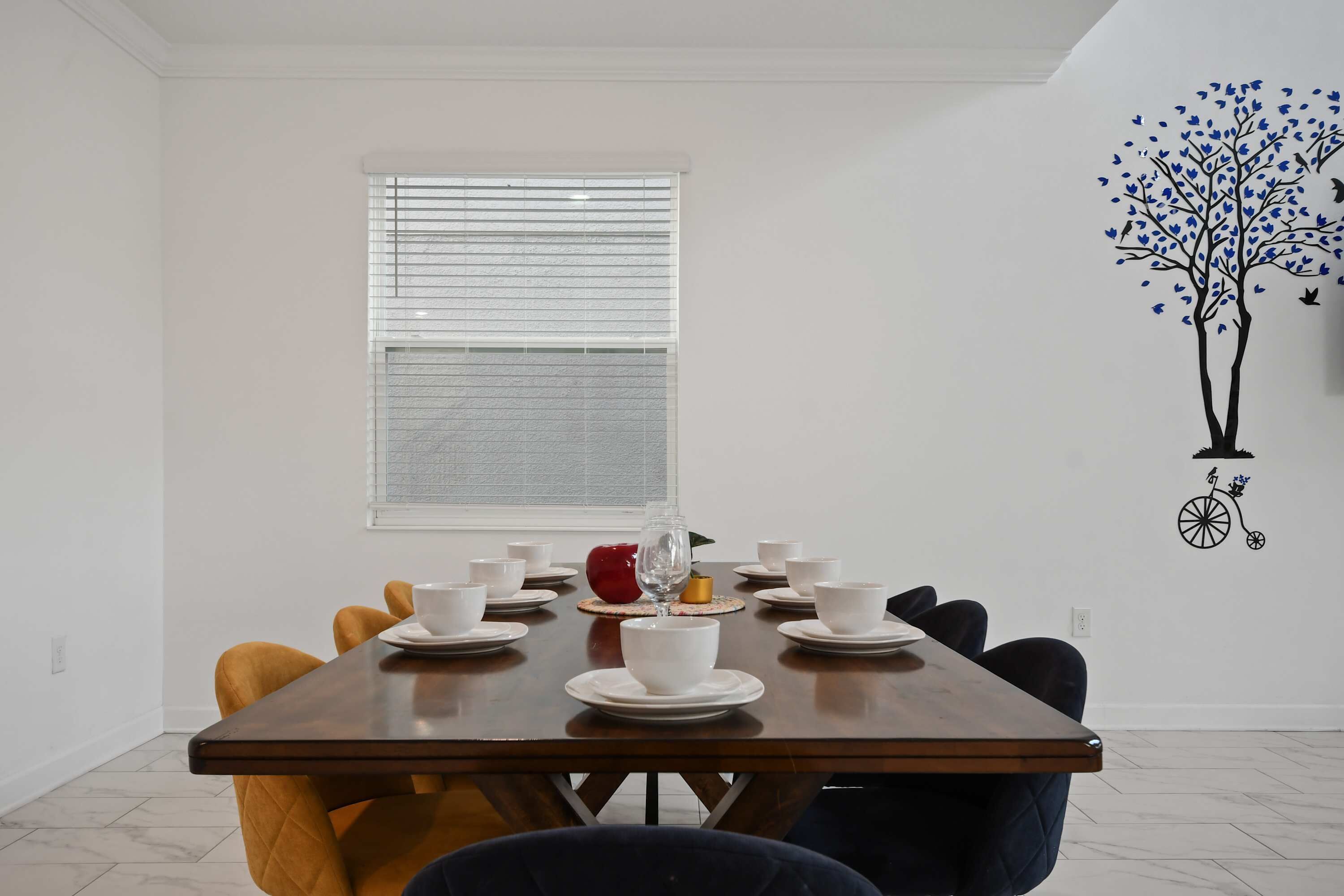 Un coin repas avec une table en bois dressée pour quatre personnes, des chaises blanches et un motif décoratif représentant un arbre avec des oiseaux sur un mur.