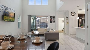 A spacious living room with a high ceiling, a dining area with a wooden table set for a meal, and a kitchen area with modern appliances. - Luxurious Resort Home Near Disney & Shopping! (Kissimmee)
