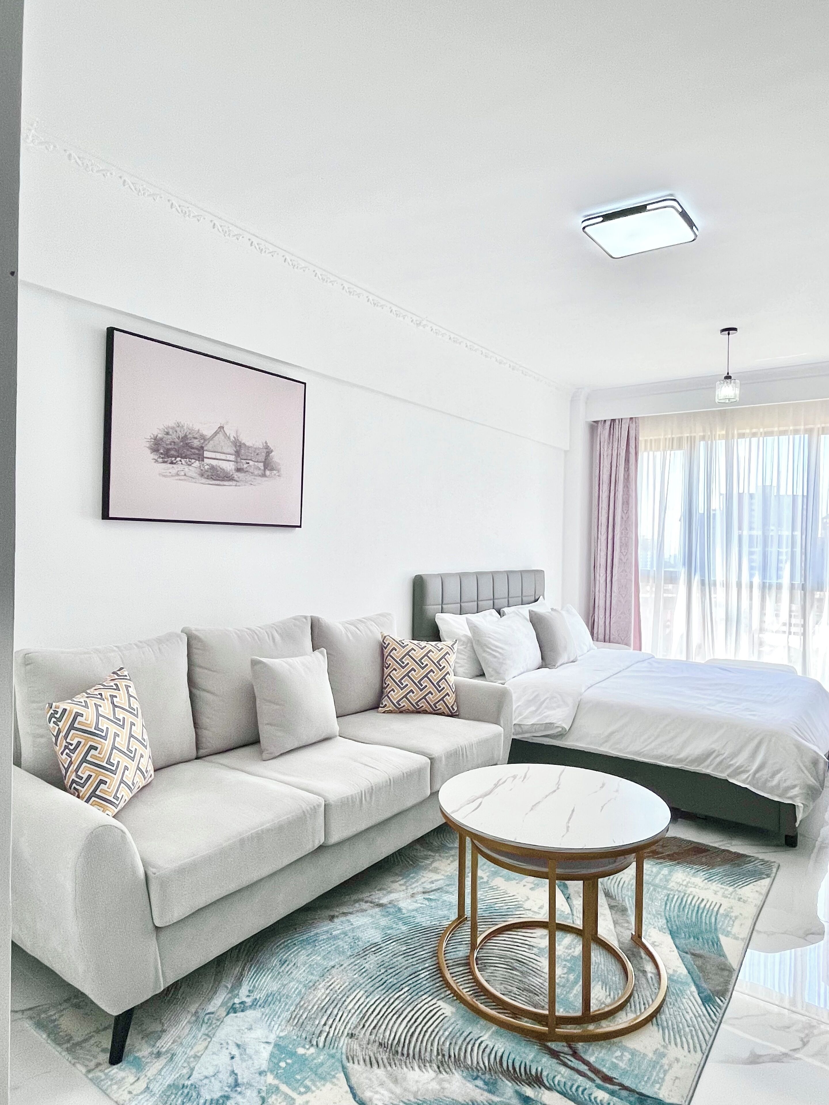 Quarto de hotel moderno com sofá cinzento, mesa de centro redonda, cama com roupa de cama cinza e quadro na parede.