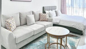 Ein modernes Hotelzimmer mit grauem Sofa, einem runden Couchtisch, einem Bett mit grauen Bettwäsche und einem gerahmten Bild an der Wand.