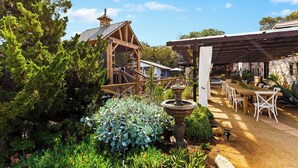 Una estructura rústica de madera con escaleras, rodeada de abundante vegetación y un comedor con sillas blancas y una mesa larga.