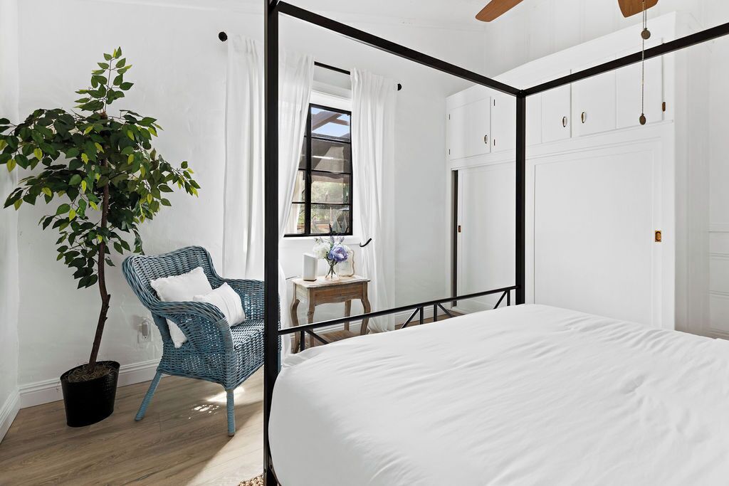 Une chambre à coucher avec un lit à baldaquin, une plante en pot, une chaise en osier et une petite table sur laquelle est posé un vase.