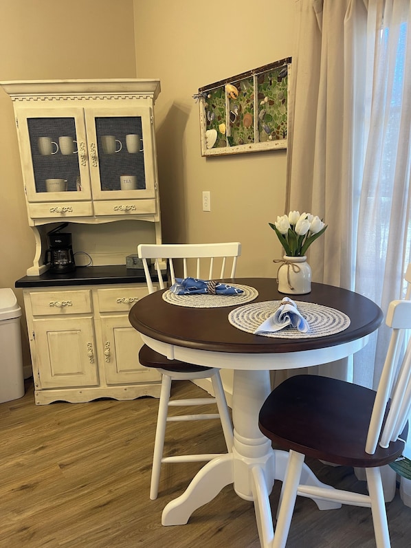 A dining area with a round table, white chairs, a coffee maker, and a cabinet with glass doors.