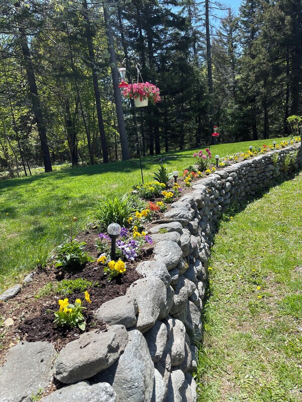 A garden with a stone retaining wall, various flowers, and a hanging planter with pink flowers.