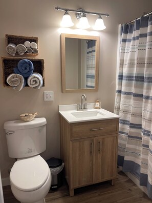 A bathroom with a white toilet, wooden cabinets, a sink with a mirror, and a striped shower curtain. - “Low Tide” cozy studio for two!  (Tremont)