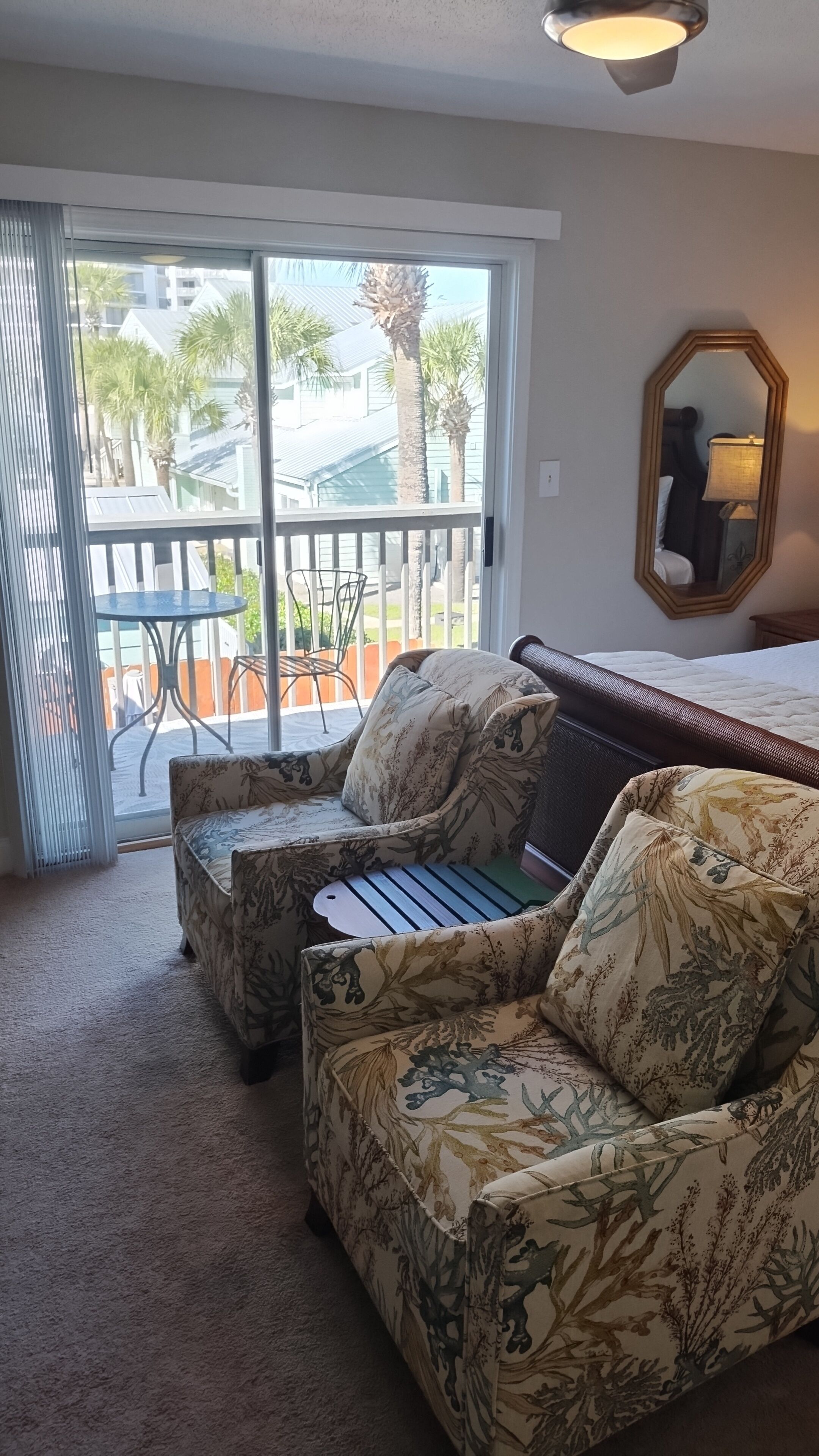 A room with a sofa and a chair, a bed, a mirror, and a sliding glass door leading to an outdoor area with a table and chairs.
