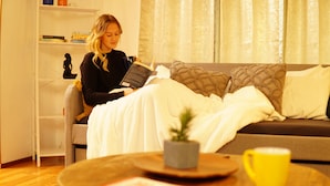 Eine Person liest ein Buch auf einem Sofa, neben einem Beistelltisch stehen eine Tasse Kaffee und ein Buch.
