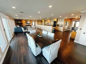A spacious living area with a dining table, white chairs, and a kitchen in the background.