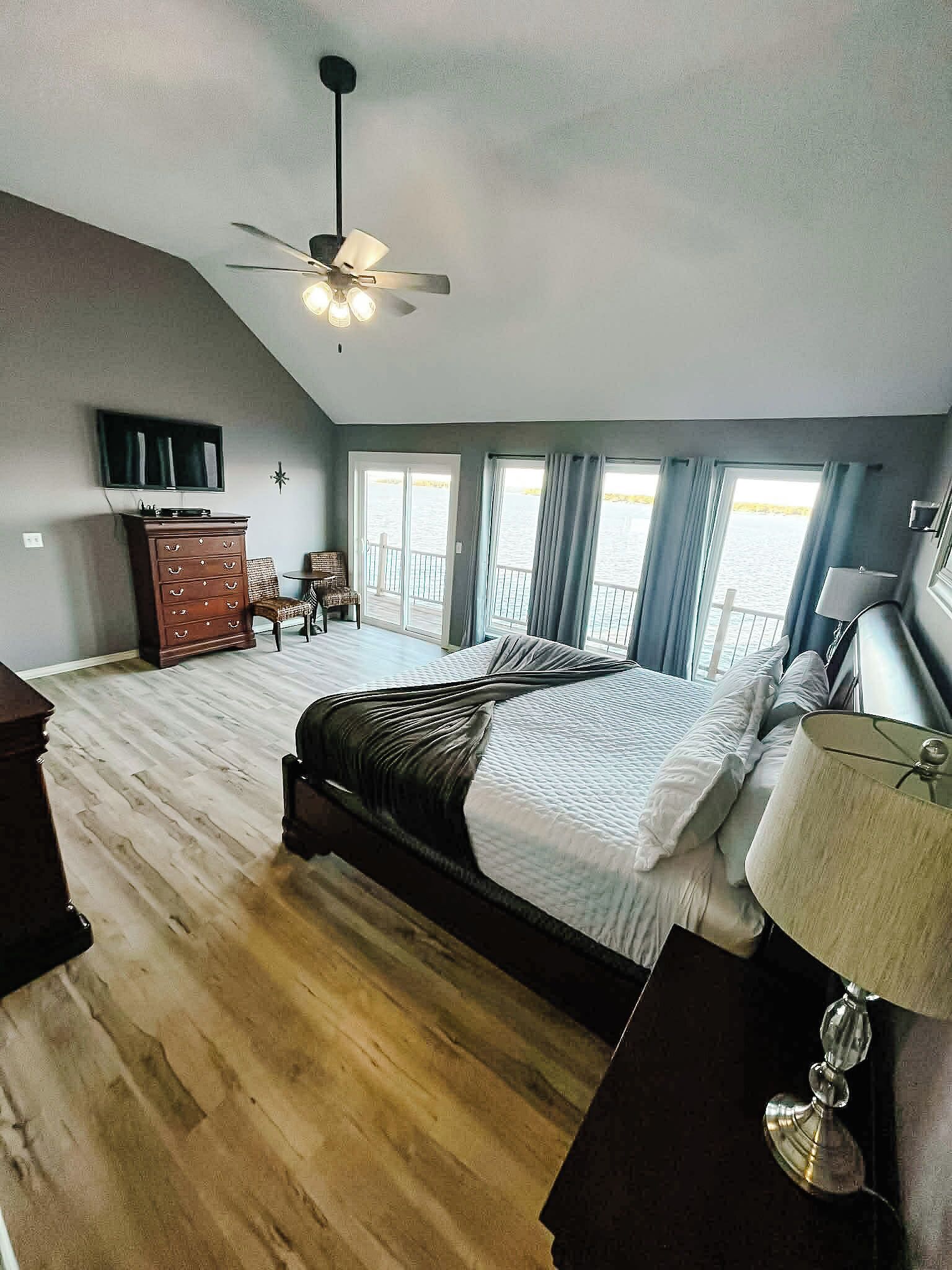 A bedroom with a bed, a dresser, a TV, a ceiling fan, and a view of the water.