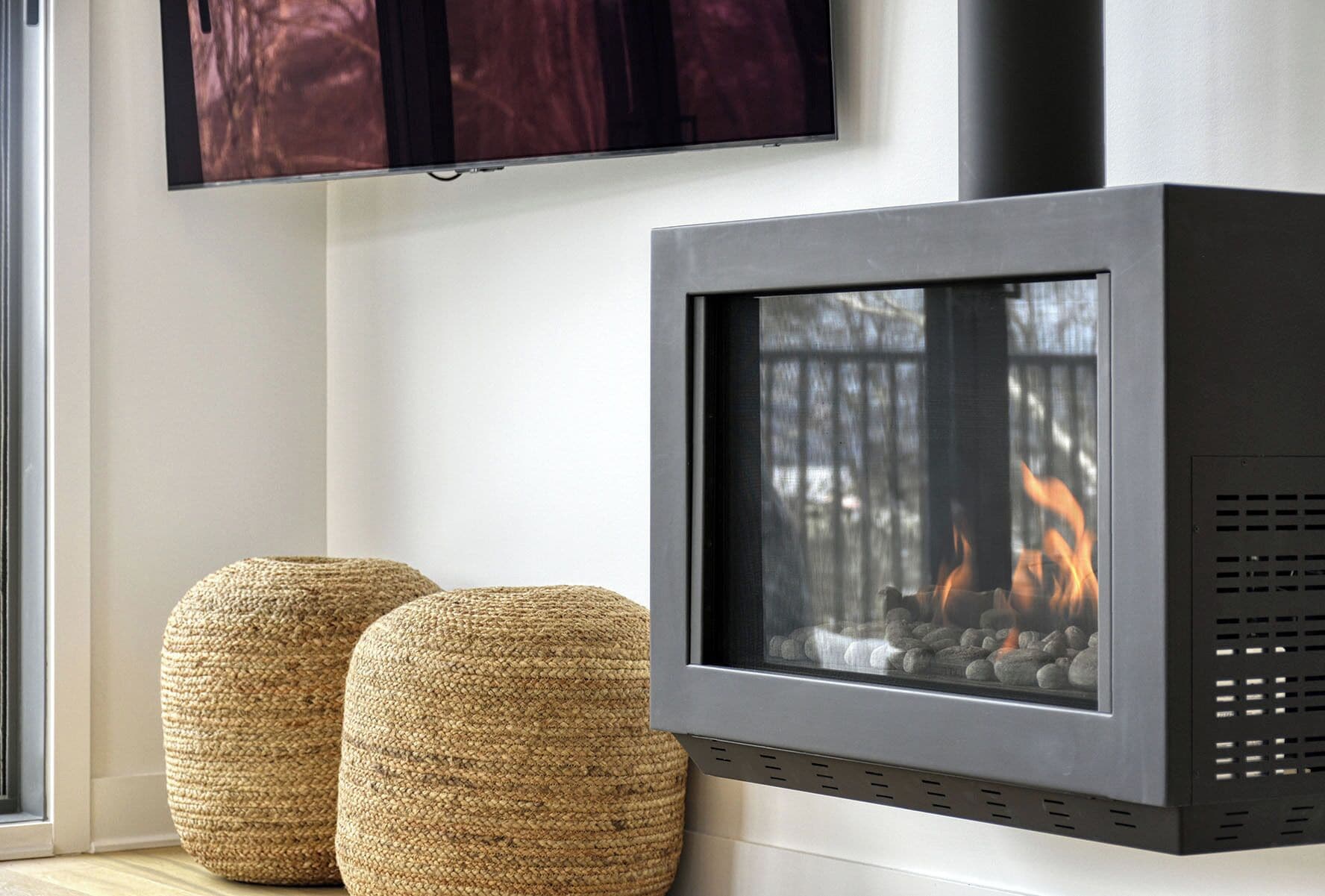 A modern fireplace with a glass front, two woven poufs, and a TV mounted above.