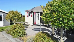 A small, single-story building with a gray roof and red and white striped awning. There is a picnic table and a small garden area with various plants. - Chalet Florence 5 , 6- Persoons met Veel Privacy; Huisdiervrij,ruime Tuin (Serooskerke)