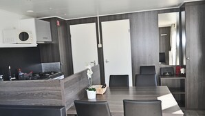 A modern kitchen with dark wood paneling, a built-in stove, and a dining area with a long table and chairs. - Chalet Florence 5 , 6- Persoons met Veel Privacy; Huisdiervrij,ruime Tuin (Serooskerke)