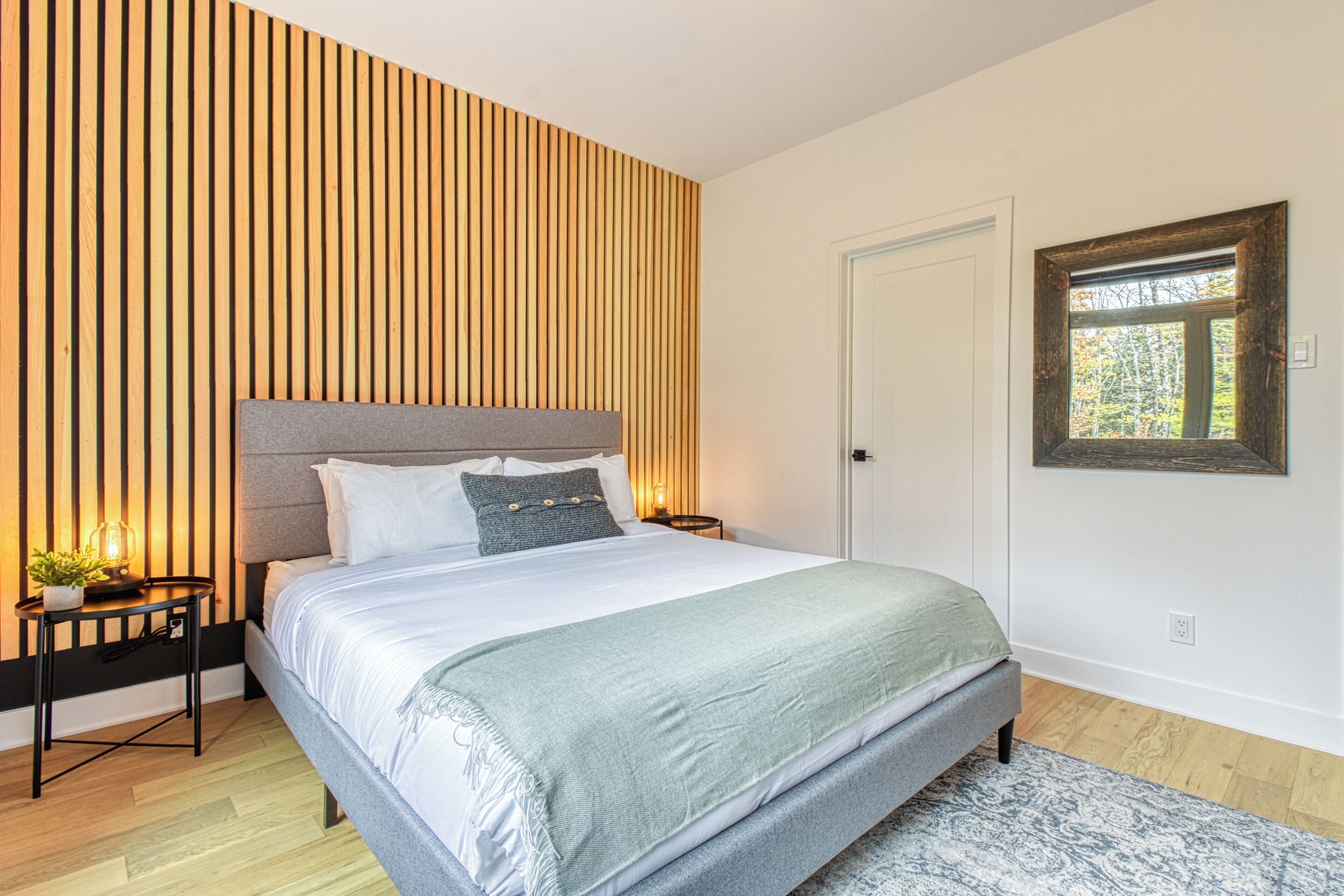 A bedroom with a bed, bedside tables, a nightstand, a wall-mounted mirror, and a window.