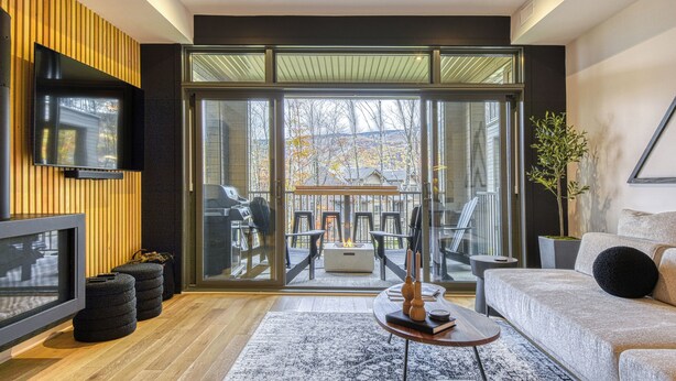 A modern living room with a flat-screen TV, a sofa, a coffee table, and a large sliding glass door leading to an outdoor seating area. - 102 Verbier Building 7 - Alpinhaus (Mont-Tremblant)