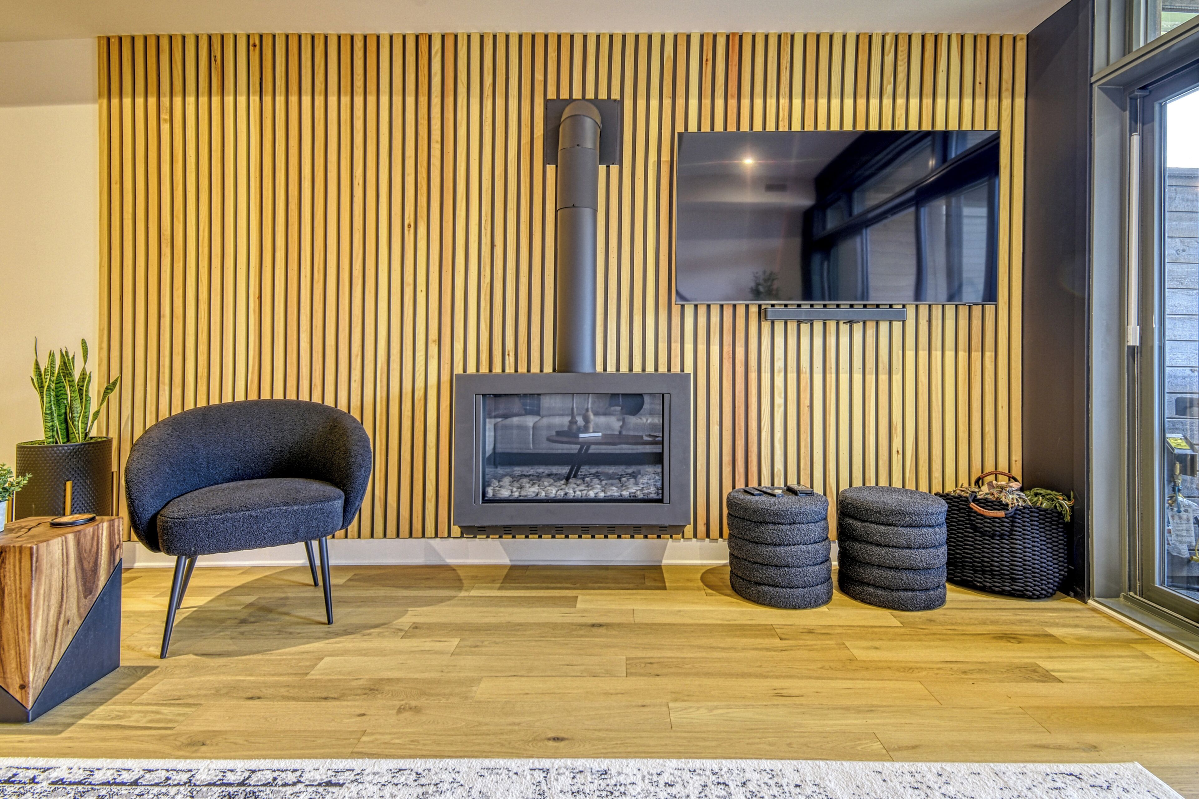 A modern living room with a wood-paneled wall, a fireplace, a comfortable chair, and a small table with a plant.
