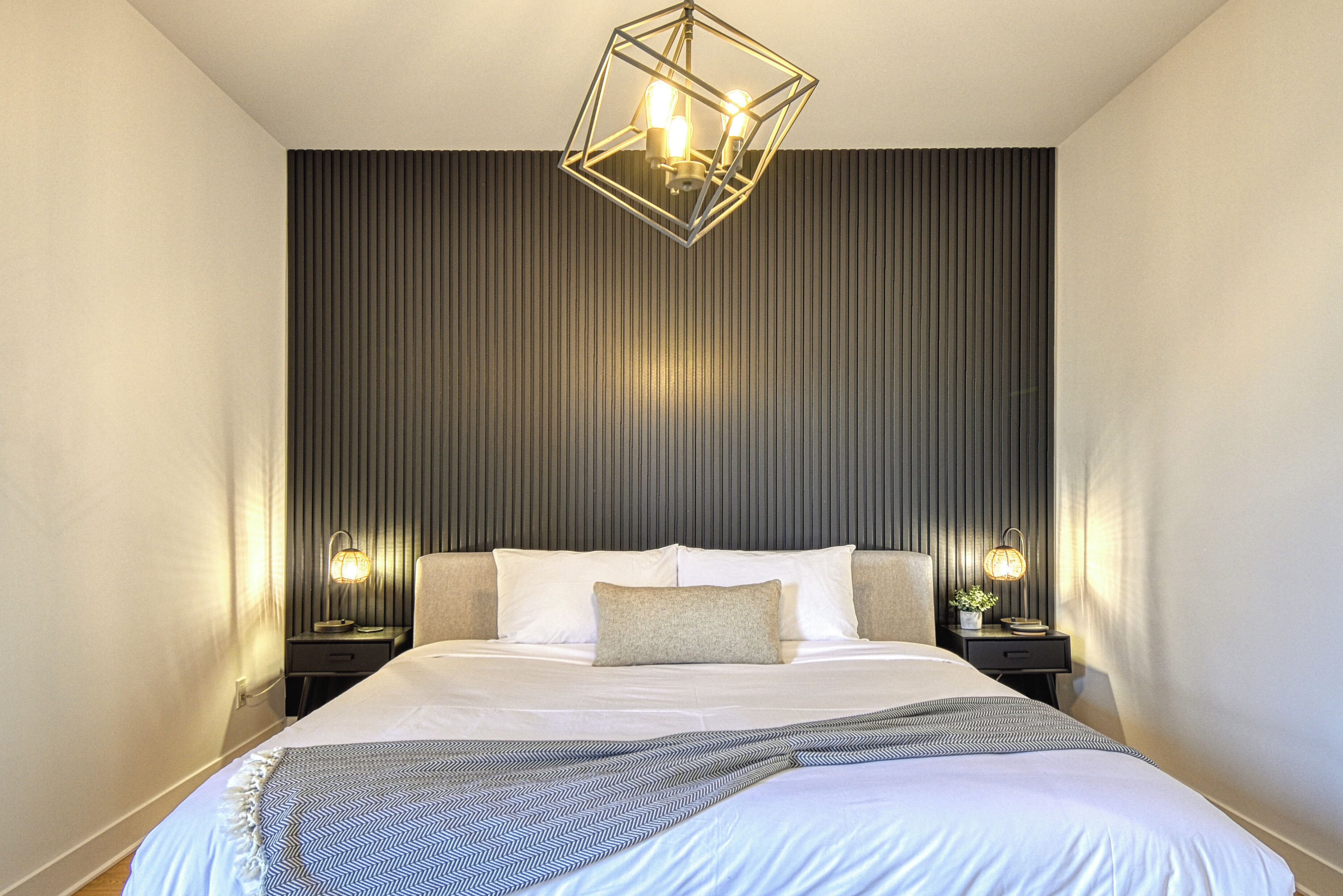 A modern bedroom with a large bed, two bedside tables, a geometric ceiling light, and a textured wall.