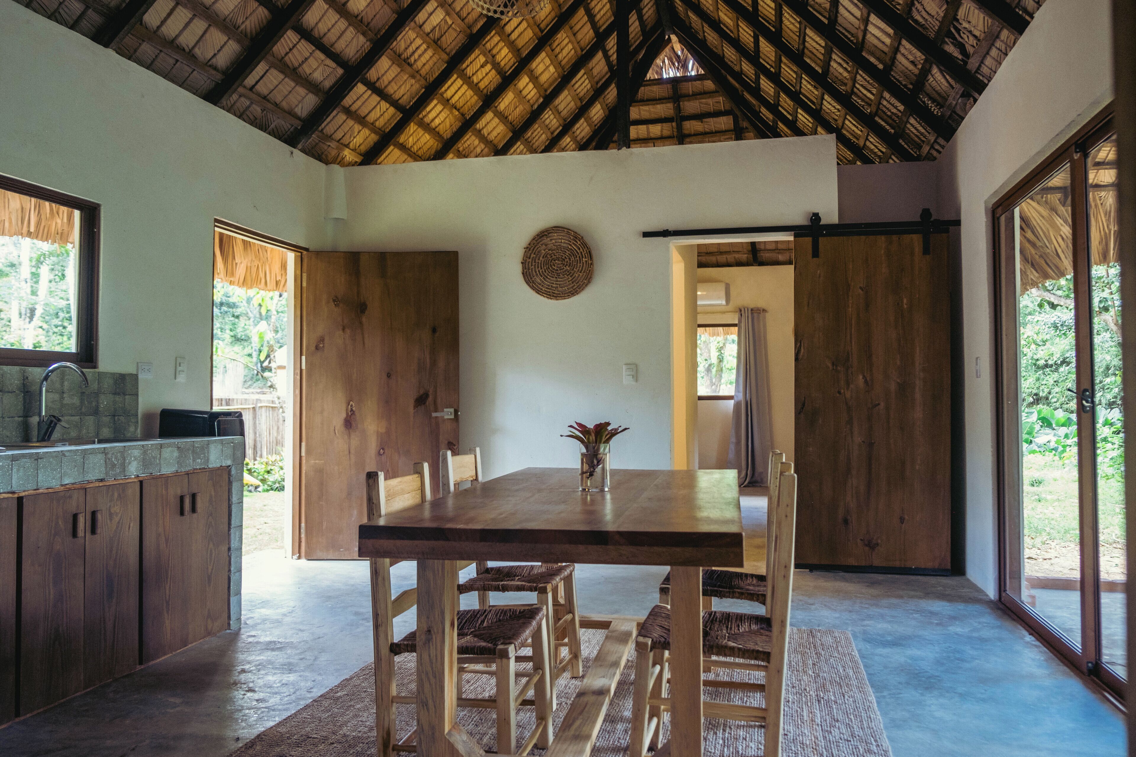 A spacious dining area with a large wooden table and chairs, a kitchen area with wooden cabinets, and large windows offering views of the outdoors.