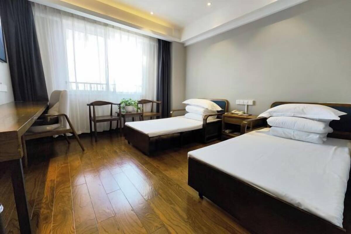A hotel room with a wooden floor, a bed, a desk, a chair, and a small table with a plant.
