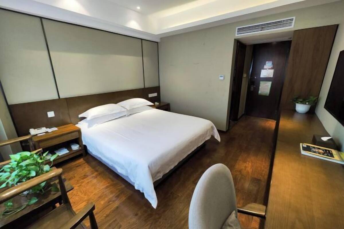 A hotel room with a large bed, wooden flooring, a desk with a chair, a nightstand, and a potted plant.