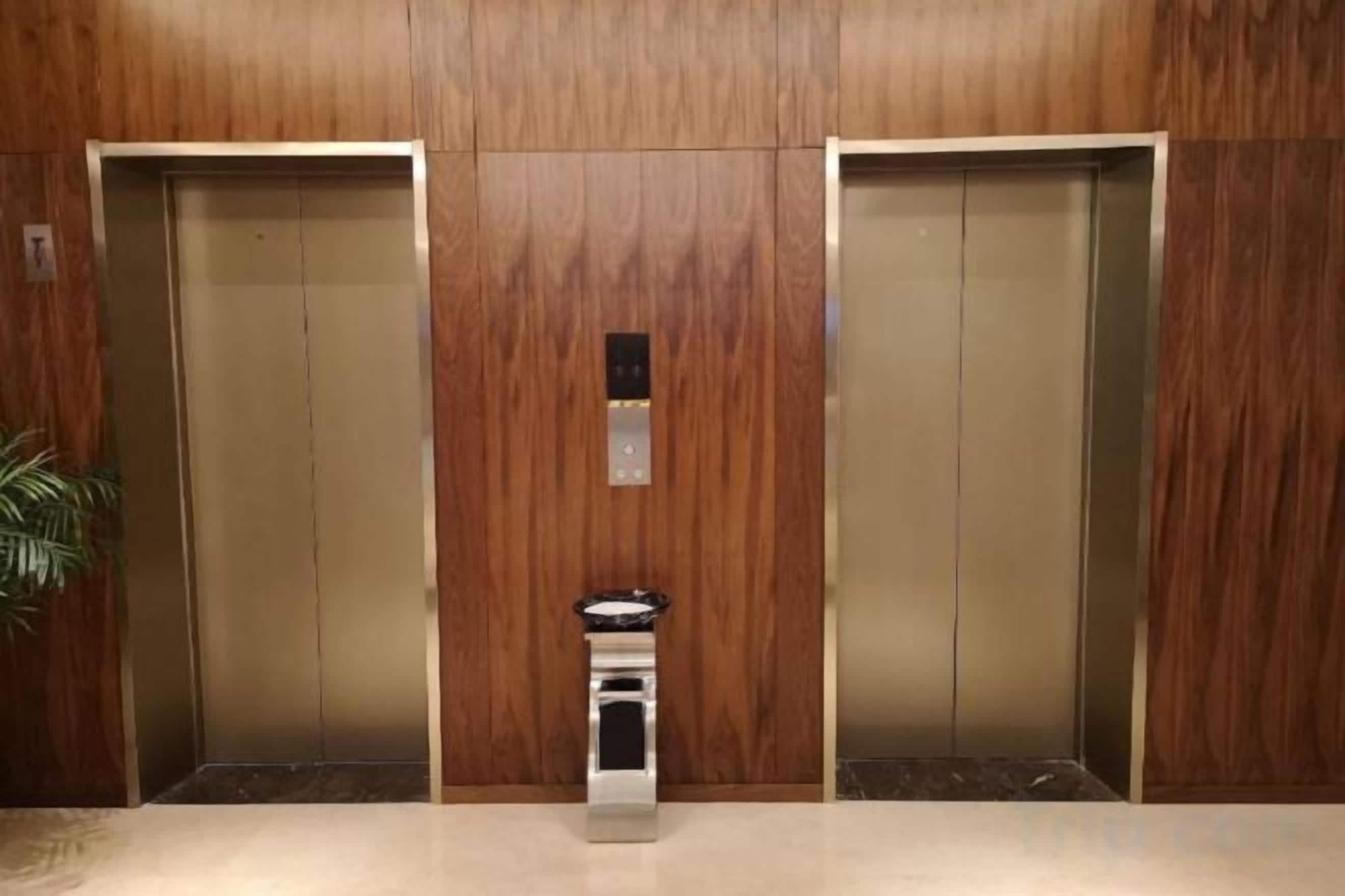 Two elevators with a central control panel between them, set against a wooden wall.