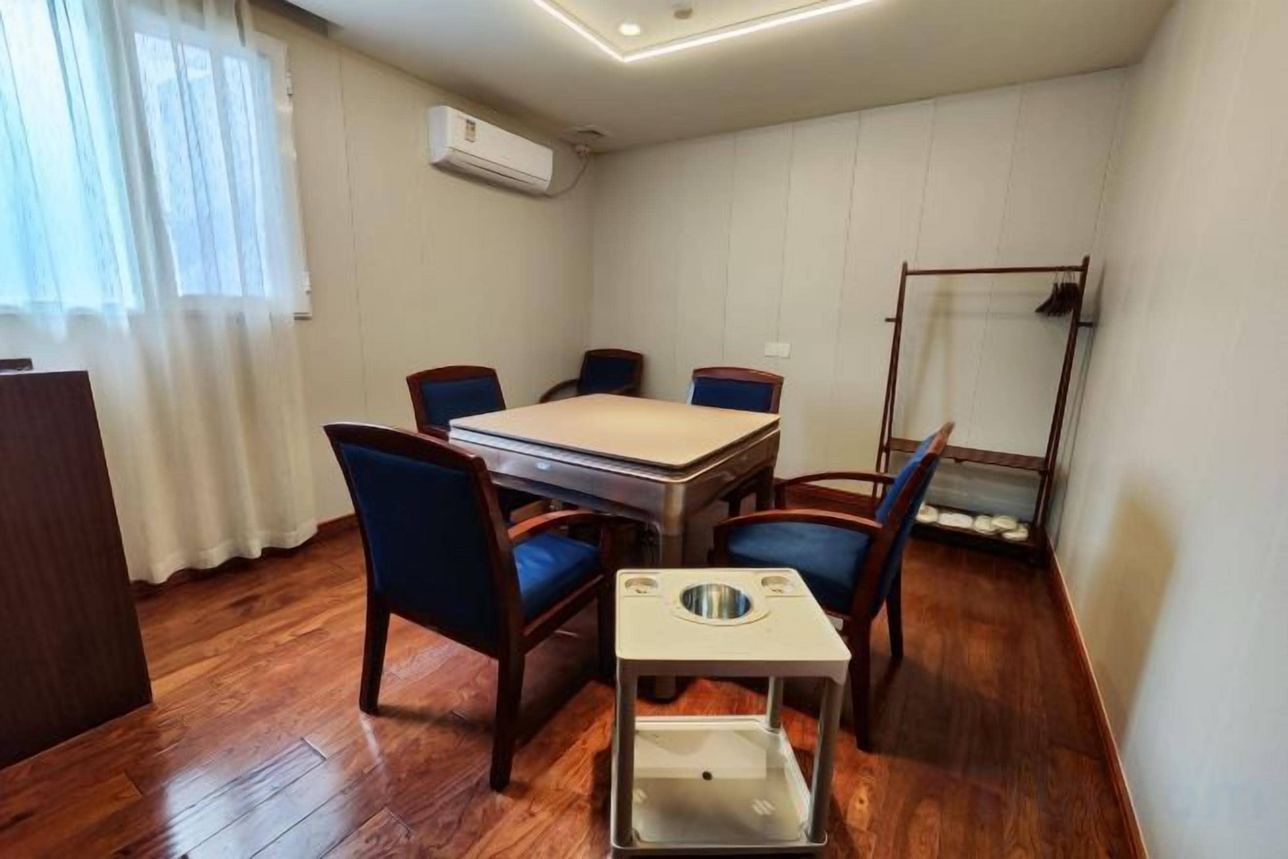 Une pièce avec un plancher en bois, une table à manger avec des chaises, une petite table avec un récipient, et une fenêtre avec des rideaux.