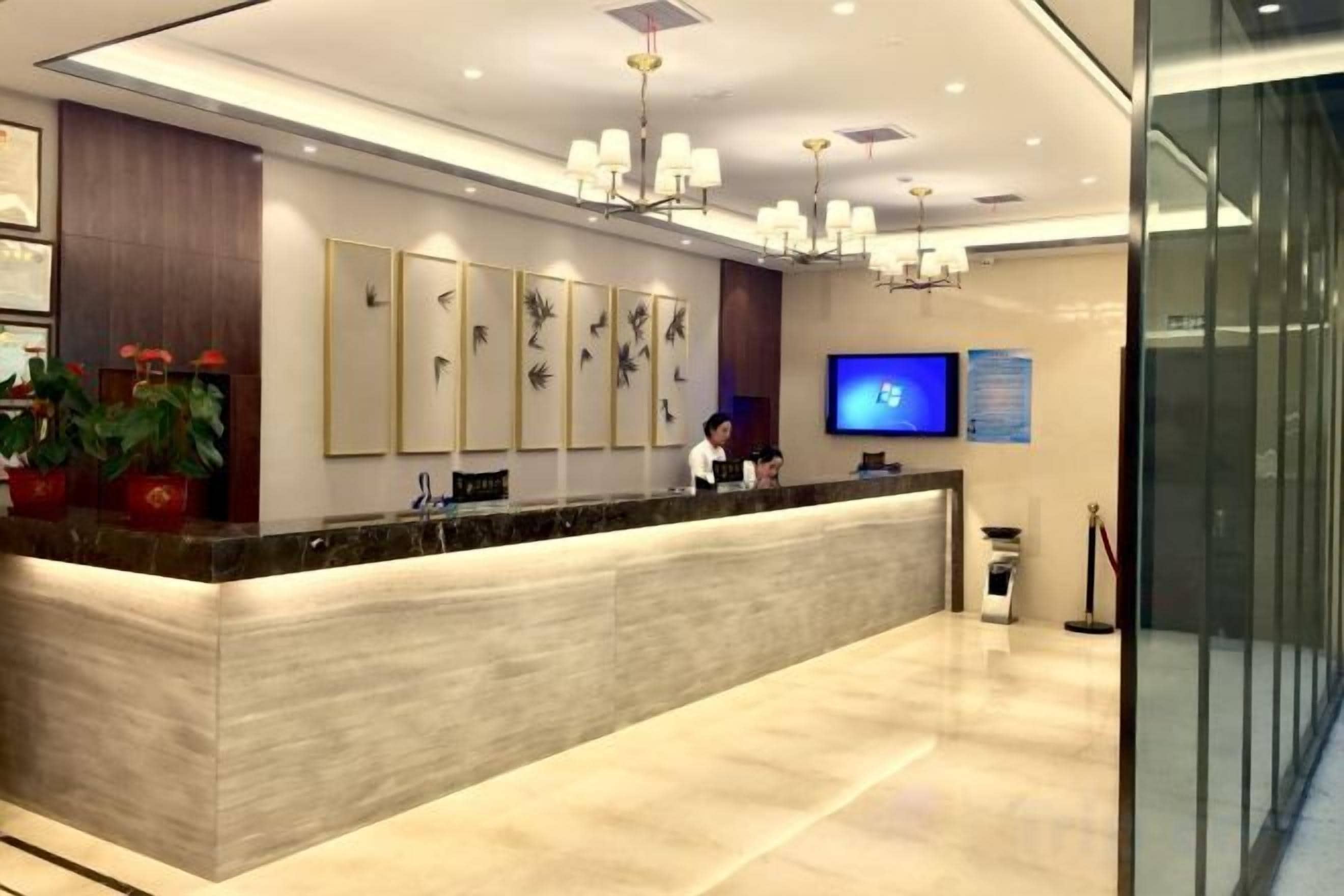 A hotel reception area with a marble counter, wall-mounted artwork, a television, and a chandelier.